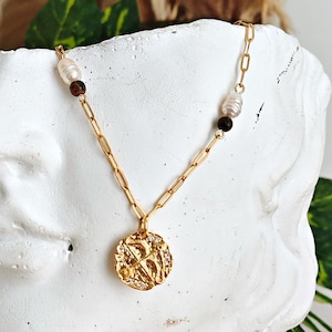 May include: A gold necklace with a circular pendant and a chain. The necklace features small pearls and dark brown beads. The pendant has a textured surface with an embossed design. The necklace is displayed on a white bust.