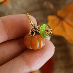 Può includere: Una collana con catena dorata con un piccolo ciondolo a forma di zucca arancione e un ciondolo a forma di foglia in vetro verde.