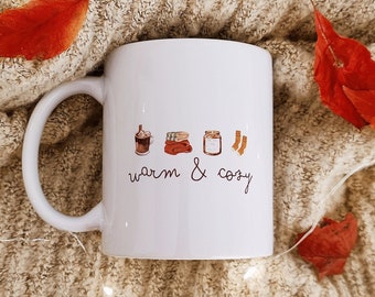 May include: White ceramic mug with a black handle. The mug has a graphic of a cup of hot chocolate, a stack of books, a candle, and a pair of socks. The text "warm & cozy" is written below the graphic.