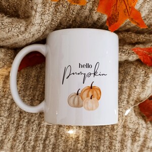 Op de afbeelding: Witte keramische mok met een zwart handgeschreven ontwerp dat "hello pumpkin" zegt en drie geillustreerde pompoenen in tinten oranje en beige.