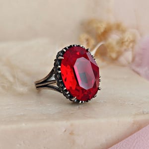 Könnte beinhalten: Ein silberfarbener Ring mit einem großen, oval geschliffenen, leuchtend roten Edelstein. Der Edelstein ist von einer detaillierten, dunklen Metalleinfassung umgeben. Der Ring hat ein geteiltes Schaftdesign. Der Ring wird auf einer hellen Oberfläche präsentiert.