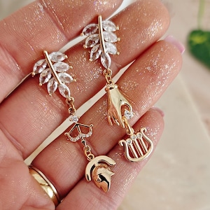 May include: A pair of gold earrings with a leaf design and dangling charms. One earring has a charm of a hand holding a lyre, and the other earring has a charm of an arrow and a helmet.