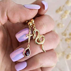 Puede incluir: Pendientes colgantes de oro con un serpiente, una cruz y un dije de piedra roja.