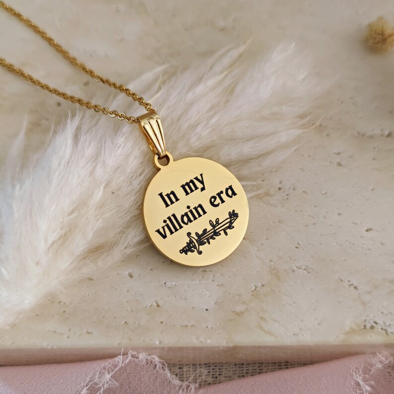 May include: Gold necklace with a round pendant that says "In my villain era" with a sword and leaves design.
