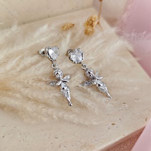 May include: A pair of silver-toned earrings featuring a heart-shaped gemstone at the top and a small cherub figure below. The cherubs have outstretched wings and are in a praying pose. The earrings are displayed on a textured, light-colored surface.
