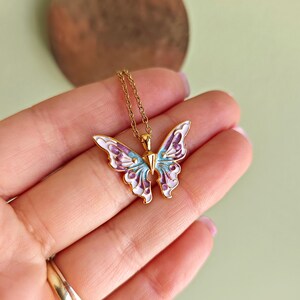 May include: A gold-colored butterfly pendant necklace. The butterfly has white, purple, and light blue enamel detailing. The necklace is on a gold chain.