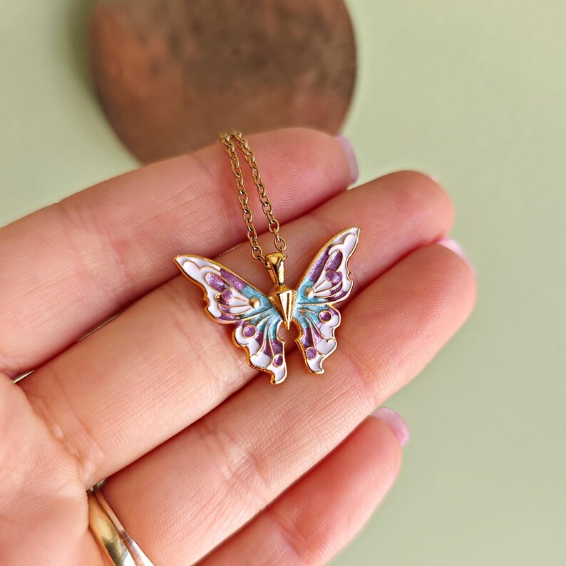 May include: A gold-colored butterfly pendant necklace. The butterfly has white, purple, and light blue enamel detailing. The necklace is on a gold chain.