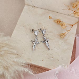 May include: A pair of silver-colored angel earrings. Each earring features a heart-shaped stud with a clear stone, connected to a detailed angel pendant with outstretched wings. The earrings are displayed on a light beige surface.