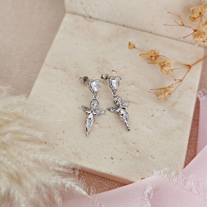 May include: A pair of silver-colored angel earrings. Each earring features a heart-shaped stud with a clear stone, connected to a detailed angel pendant with outstretched wings. The earrings are displayed on a light beige surface.