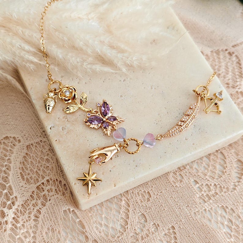 May include: A gold chain necklace with multiple charms, including a butterfly, a rose, a hand, a feather, and a star. The charms are adorned with purple and clear crystals.