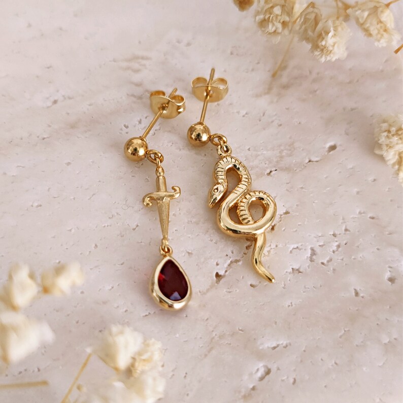 May include: Gold dangle earrings with a red gemstone, a dagger, and a snake charm.