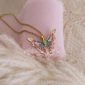 May include: A gold-colored butterfly pendant necklace. The butterfly has pink, blue, and white enamel detailing. The necklace is displayed on a pink heart-shaped cushion and white fluffy feathers.