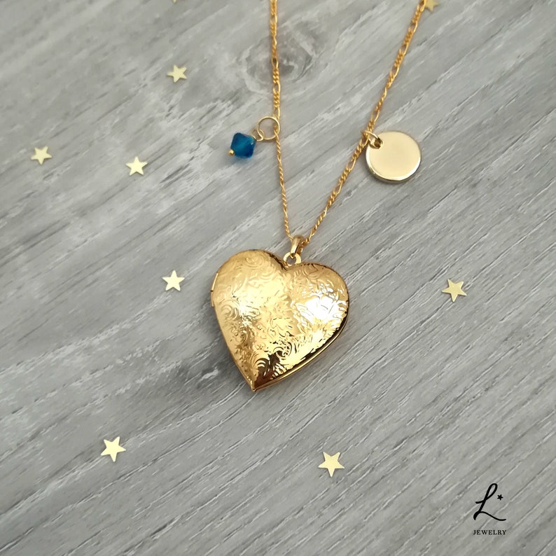 May include: Gold heart-shaped locket necklace with a blue crystal charm and a round gold charm. The necklace is on a wooden surface with gold stars.