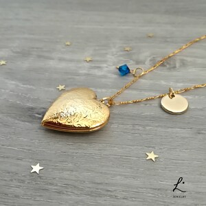 May include: Gold heart locket necklace with a small round charm and a blue crystal accent. The necklace is on a wood background with gold stars.