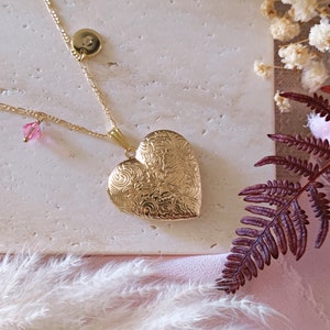 May include: A gold heart-shaped locket necklace with a delicate chain and a small pink crystal charm. The locket has an intricate floral design.