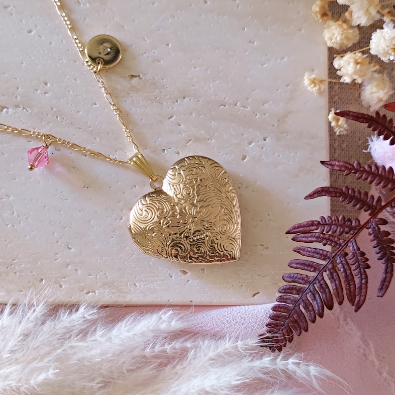 May include: A gold heart-shaped locket necklace with a delicate chain and a small pink crystal charm. The locket has an intricate floral design.