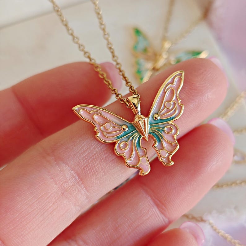 May include: A gold-colored butterfly pendant necklace with a delicate chain. The butterfly has pink and teal enamel detailing. The necklace is held in a hand, with another butterfly necklace visible in the background.
