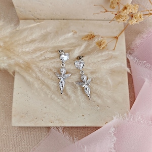 May include: A pair of silver-toned dangle earrings. Each earring features a heart-shaped stud, a small cherub figure with wings, and a dangling design. The earrings are displayed on a neutral-toned surface with dried flowers and pink fabric.