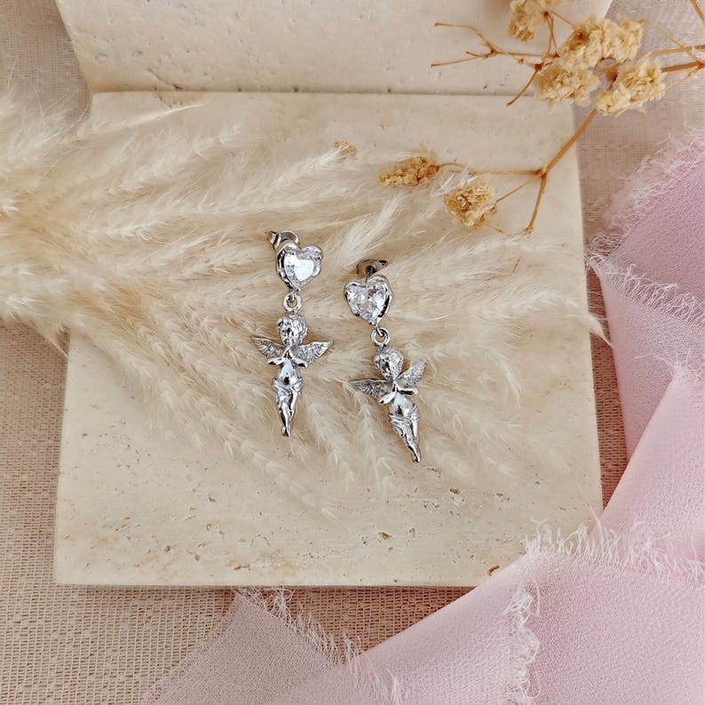 May include: A pair of silver-toned dangle earrings. Each earring features a heart-shaped stud, a small cherub figure with wings, and a dangling design. The earrings are displayed on a neutral-toned surface with dried flowers and pink fabric.