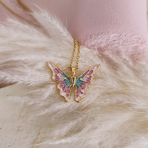 May include: A gold-colored butterfly pendant necklace. The butterfly has pink, blue, and purple enamel detailing. The necklace is displayed on a bed of white feathers and a pink background.