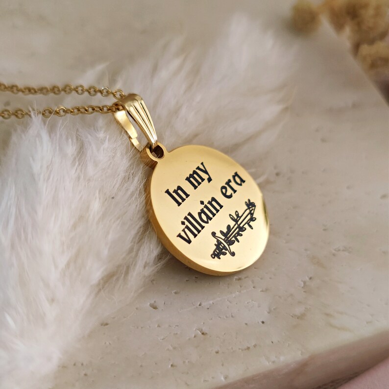 May include: Gold necklace with a round pendant that says "In my villain era" with a sword and leaves design.