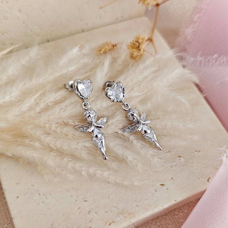 May include: Silver-toned dangle earrings featuring a heart-shaped stud and a cherub pendant. The cherub has detailed wings and is holding its hands together. The earrings are displayed on a neutral surface with a soft, feathery texture.