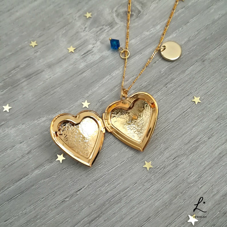 May include: Gold heart-shaped locket necklace with a blue crystal charm and a round pendant. The locket is open, revealing a detailed engraved design.