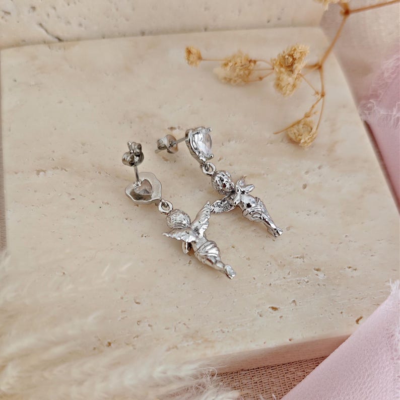 May include: Silver-toned dangle earrings featuring a heart-shaped stud and a dangling cherub design. The earrings have a small, clear gemstone and are displayed on a neutral surface.