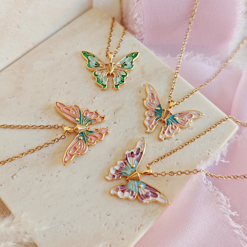 May include: Four gold-colored butterfly pendant necklaces. Each butterfly pendant features a different color scheme, including green, pink, and purple, with gold-colored chain. The necklaces are displayed on a light-colored, textured surface with a pink fabric background.