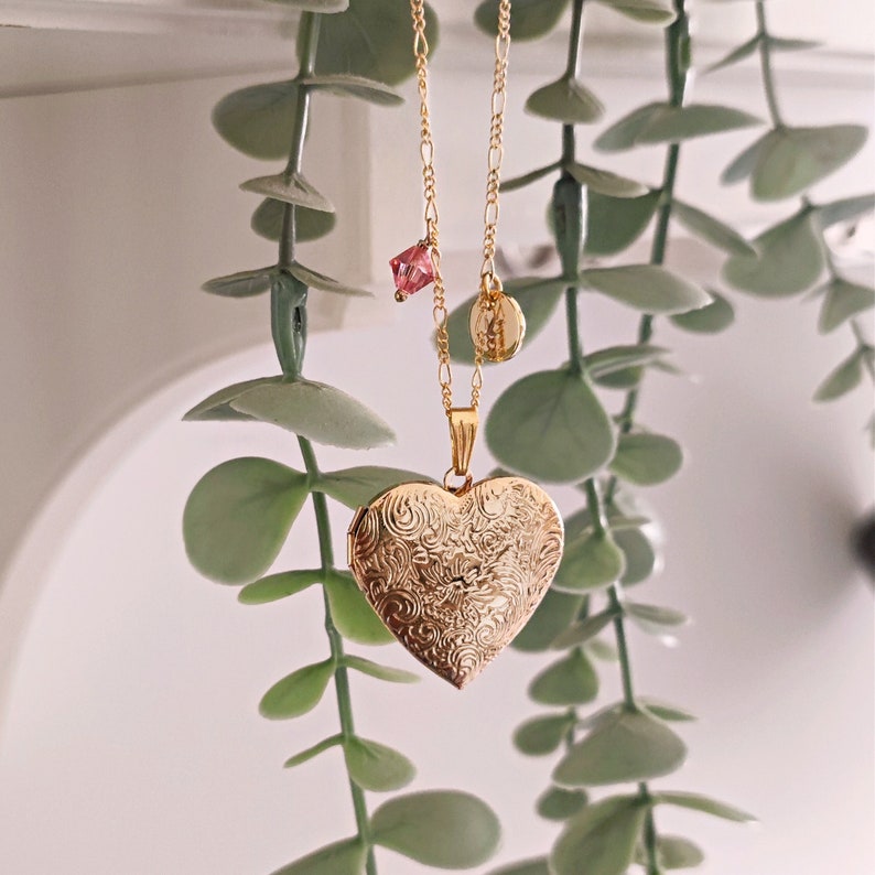 May include: A gold heart-shaped locket necklace with a delicate chain and a small pink crystal charm. The locket has a detailed floral pattern.