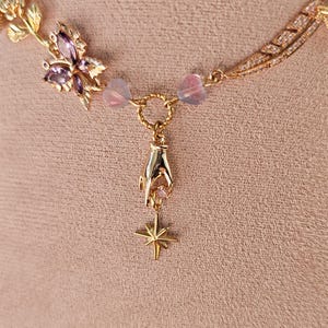 May include: Gold necklace with a delicate chain, a butterfly charm with pink and purple stones, and a hand charm holding a star pendant.