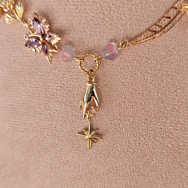 May include: Gold necklace with a delicate chain, a butterfly charm with pink and purple stones, and a hand charm holding a star pendant.