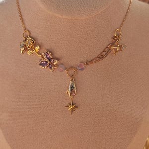 May include: A gold necklace with a delicate chain and multiple charms, including a rose, a butterfly with purple gemstones, a hand, and a star. The charms are connected by small beads.
