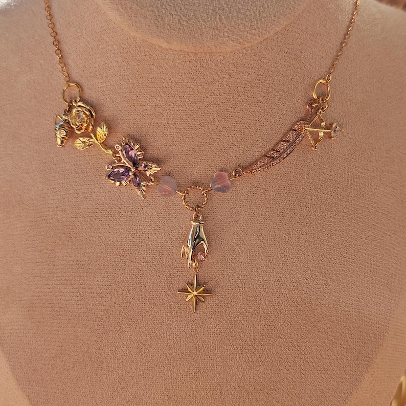 May include: A gold necklace with a delicate chain and multiple charms, including a rose, a butterfly with purple gemstones, a hand, and a star. The charms are connected by small beads.