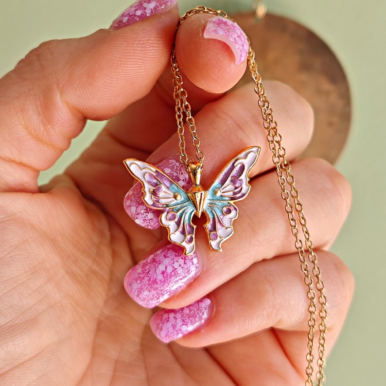 May include: A gold-colored butterfly pendant necklace. The butterfly has white, purple, and light blue enamel detailing. The necklace is held in a hand with pink nail polish.