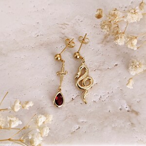 May include: A pair of gold earrings with a dangling red gemstone and a snake design. One earring features a small dagger charm.