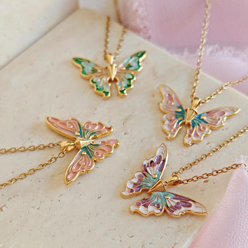 May include: Four butterfly pendant necklaces with gold chains. Each pendant features a butterfly design with colorful enamel wings in shades of pink, blue, green, and purple. The necklaces are displayed on a light-colored surface.
