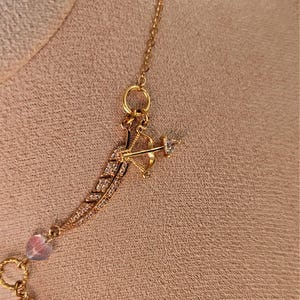 May include: A gold necklace with a feather charm and a small arrow charm. The charms are decorated with clear crystals.