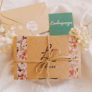 May include: A brown jewelry box tied with twine, adorned with floral patterned tape. The box is accompanied by a beige envelope, a small teal card with script, and a sticker. The box has the word "JEWELRY" printed on it.