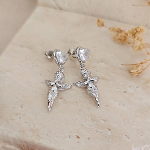 May include: A pair of silver-toned earrings featuring a heart-shaped stud and a dangling cherub design. The cherubs have outstretched arms and detailed wings. The earrings are displayed on a neutral-toned surface.