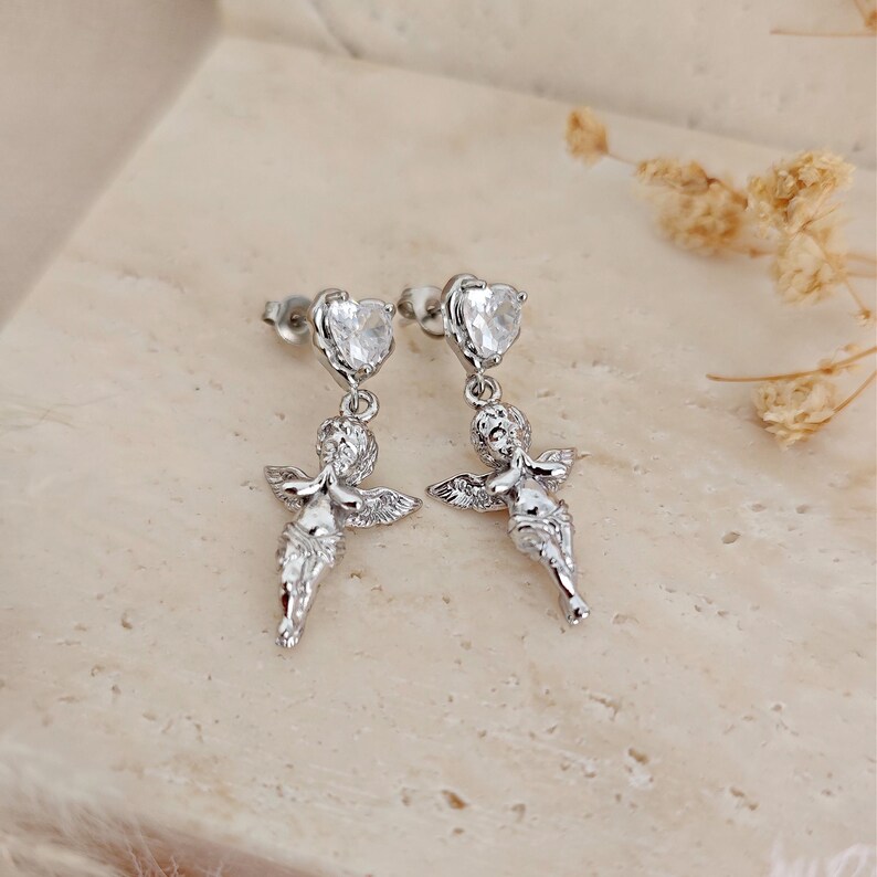 May include: A pair of silver-toned earrings featuring a heart-shaped stud and a dangling cherub design. The cherubs have outstretched arms and detailed wings. The earrings are displayed on a neutral-toned surface.
