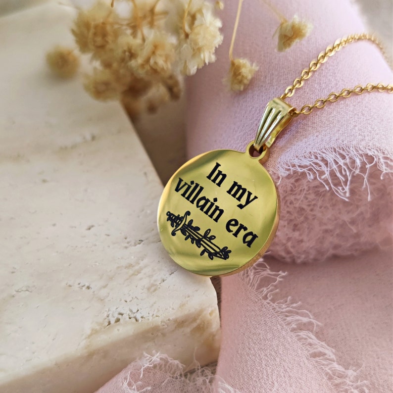 May include: Gold necklace with a round pendant that says "In my villain era" with a small sword design. The pendant is hanging on a pink fabric background.