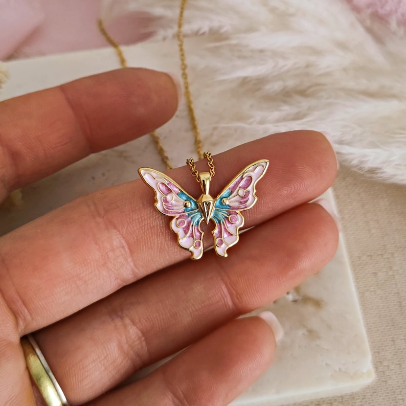 May include: A gold-colored butterfly pendant necklace. The butterfly has pink, blue, and white enamel detailing. The necklace is on a gold chain. The butterfly pendant is approximately 1.5 inches wide.