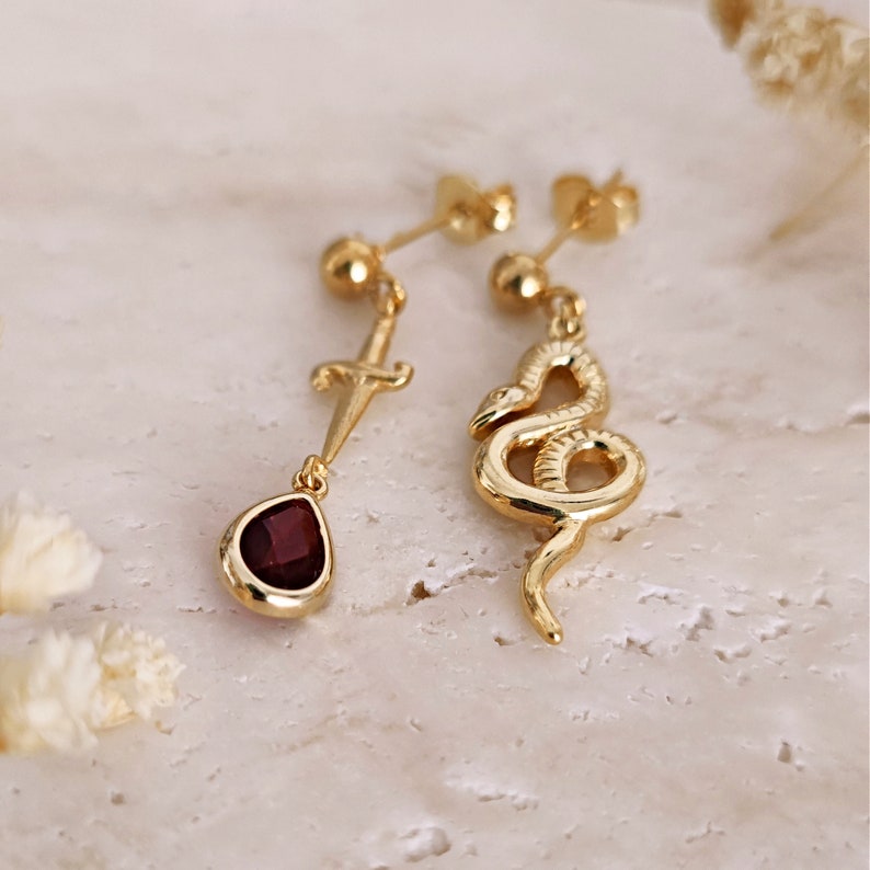 May include: Gold dangle earrings with a red gemstone, a dagger, and a snake charm.
