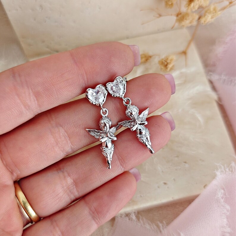 May include: Silver-toned earrings featuring a heart-shaped top with a clear stone and a cherub pendant. The cherubs have detailed wings and are in a graceful pose. The earrings are designed to dangle.