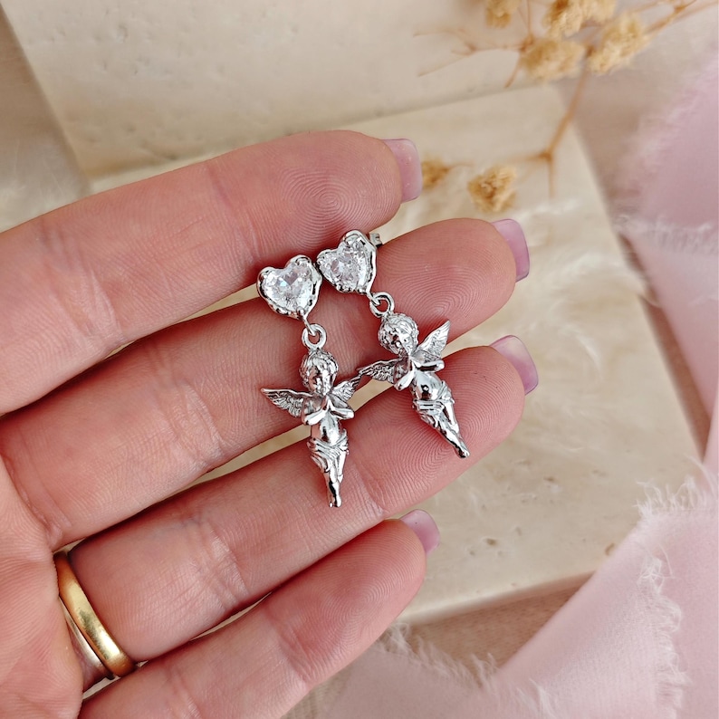 May include: A pair of silver-toned dangle earrings. Each earring features a heart-shaped stud with a clear stone, connected to a small cherub figure with outstretched wings. The cherubs are detailed with delicate features.