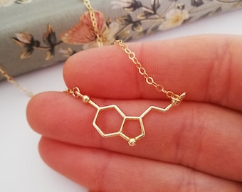 May include: A delicate gold-colored necklace featuring the molecular structure of serotonin. The pendant is a geometric design with a chain. The necklace is held in a hand, with a floral background.
