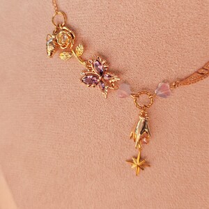 May include: A gold necklace with a delicate chain and multiple charms. The charms include a rose, a butterfly with purple gemstones, a small flower, and a star. The necklace is on a pink background.