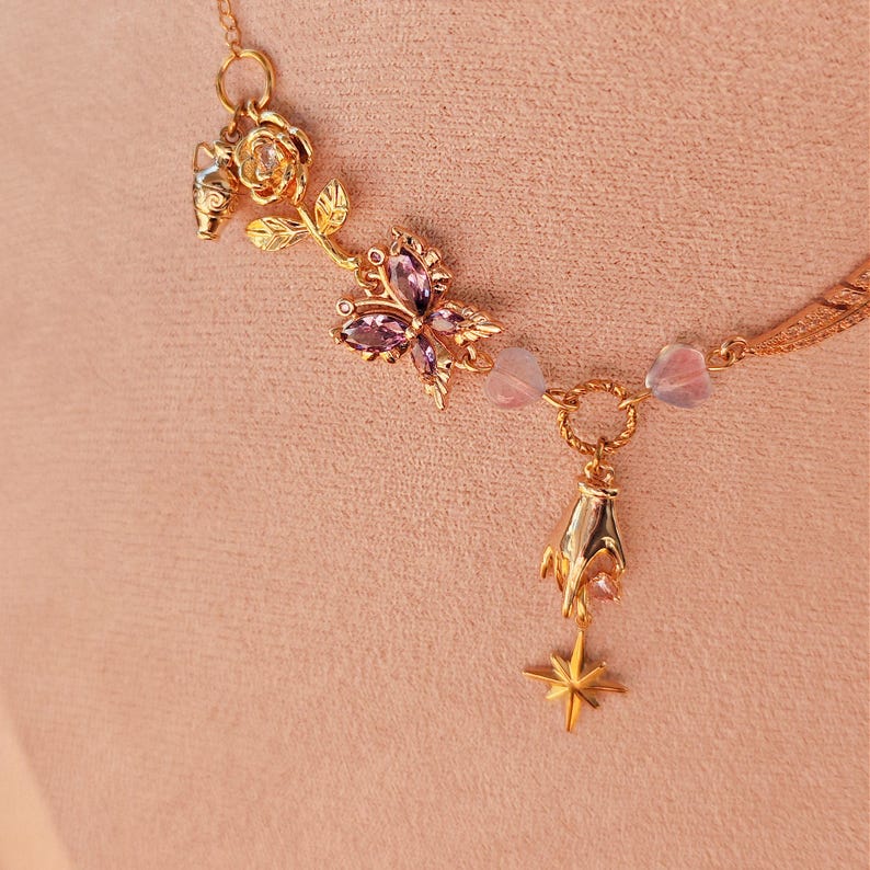 May include: A gold necklace with a delicate chain and multiple charms. The charms include a rose, a butterfly with purple gemstones, a small flower, and a star. The necklace is on a pink background.