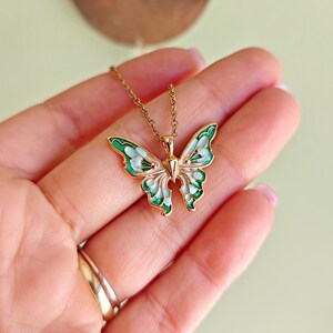 May include: A gold-colored butterfly pendant necklace. The butterfly wings are detailed with green and light blue enamel, and the necklace chain is delicate. The pendant is held in a hand, showcasing the jewelry's design and color.